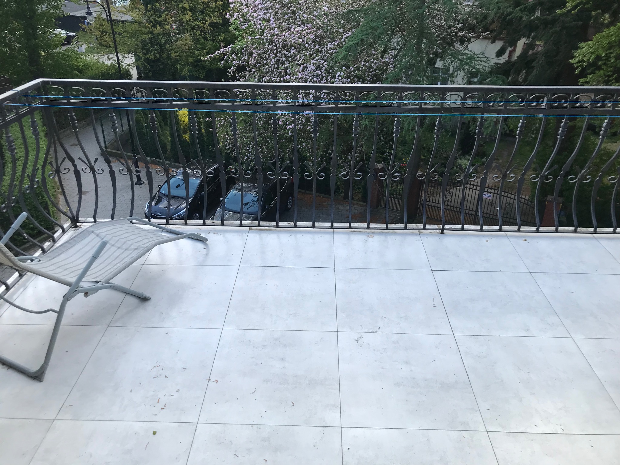 Białe płytki na balkonie z metalową balustradą, widok na zieleń i drzewa kwitnące. Krzesło plażowe w lewym rogu. Ujęcie z góry, jasne światło dzienne.