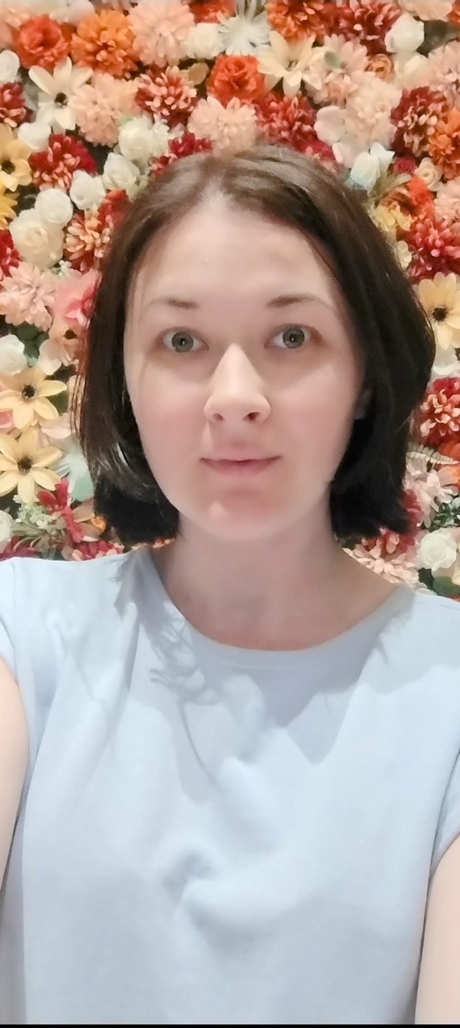 Portret kobiety z krótkimi, ciemnymi włosami na tle ściany z kwiatów w odcieniach czerwieni, pomarańczy i bieli. Ubrana w jasnoniebieską koszulkę. Delikatny makijaż, naturalny wyraz twarzy.