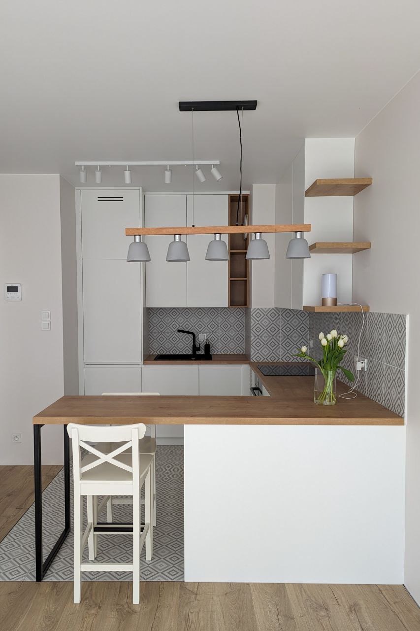 Jasna kuchnia z wyspą i drewnianym blatem. Nad wyspą lampy w szarych kloszach. W tle białe szafki i geometryczny wzór na ścianie. Na blacie wazon z białymi tulipanami.
