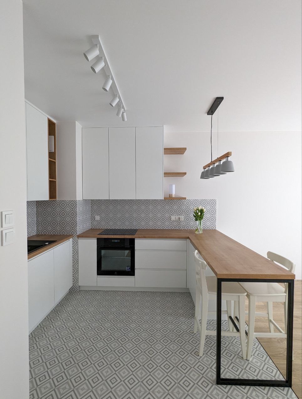 Nowoczesna kuchnia z białymi szafkami, drewnianym blatem i wzorzystą podłogą. Oświetlenie punktowe i minimalistyczny design. Wazon z kwiatami dodaje świeżości.