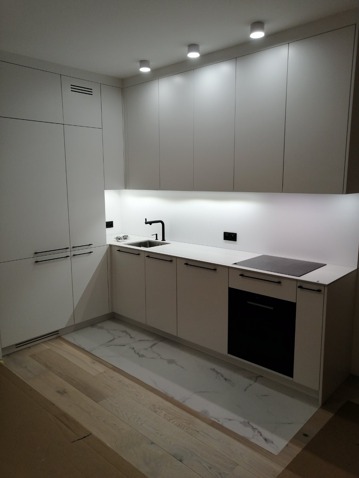 Minimalistyczna, jasna kuchnia z białymi frontami i czarnymi uchwytami. Uwagę przykuwa marmurowa podłoga i oświetlenie LED pod szafkami wiszącymi. Nowoczesny design.