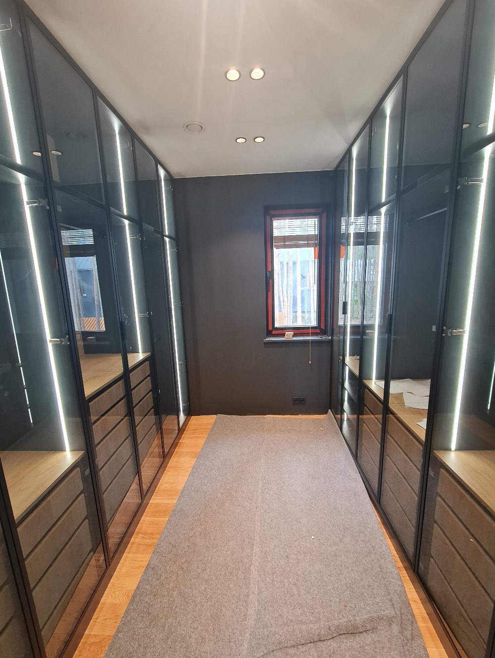 Nowoczesna garderoba z szklanymi frontami i oświetleniem LED, widok z perspektywy wejścia, szara ściana z oknem w tle, podłoga zabezpieczona szarą folią.