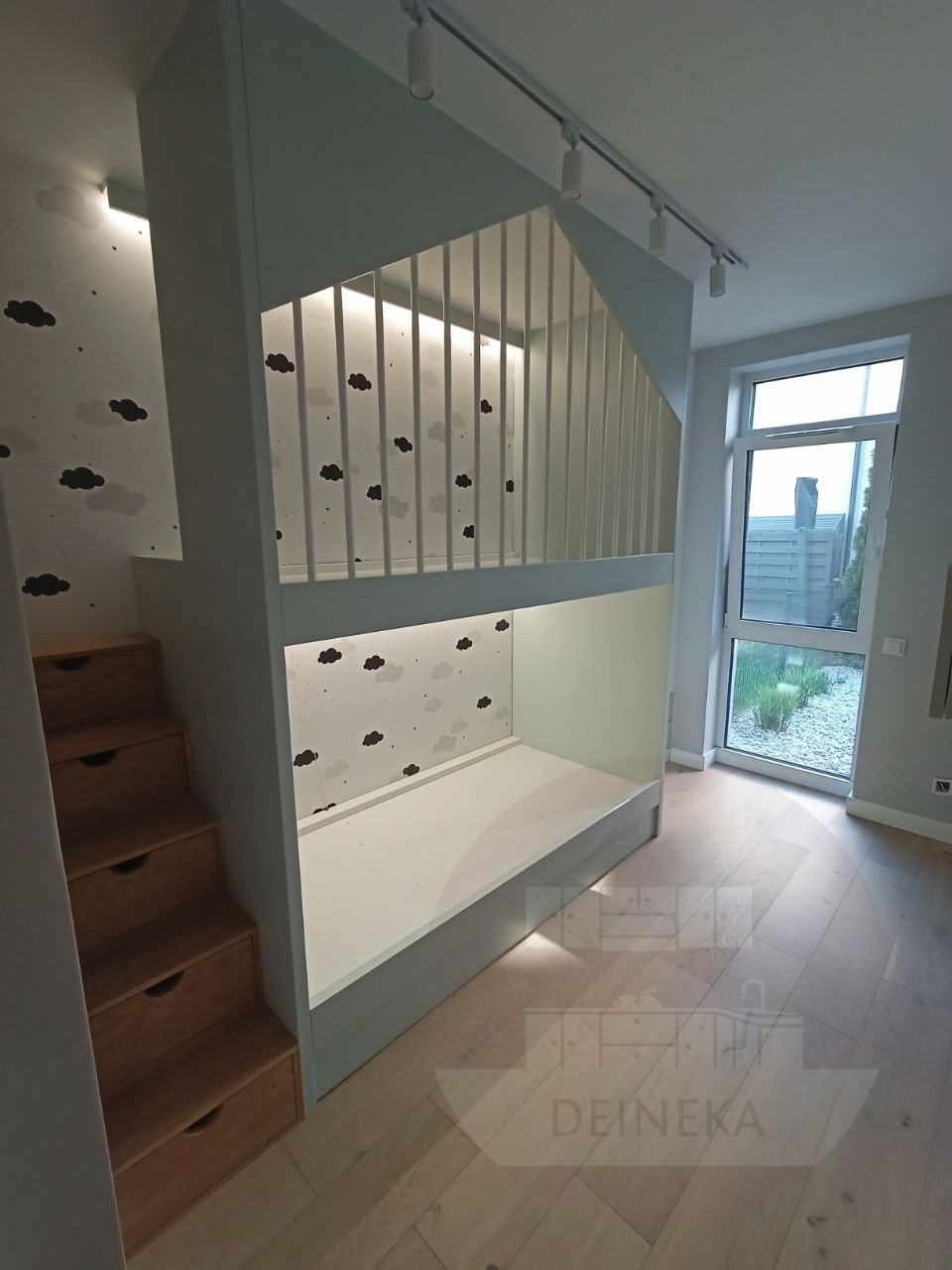 Funkcjonalne łóżko piętrowe z szufladami w schodkach, w pokoju dziecięcym z motywem chmur na ścianie. Nowoczesne oświetlenie punktowe i jasna podłoga.