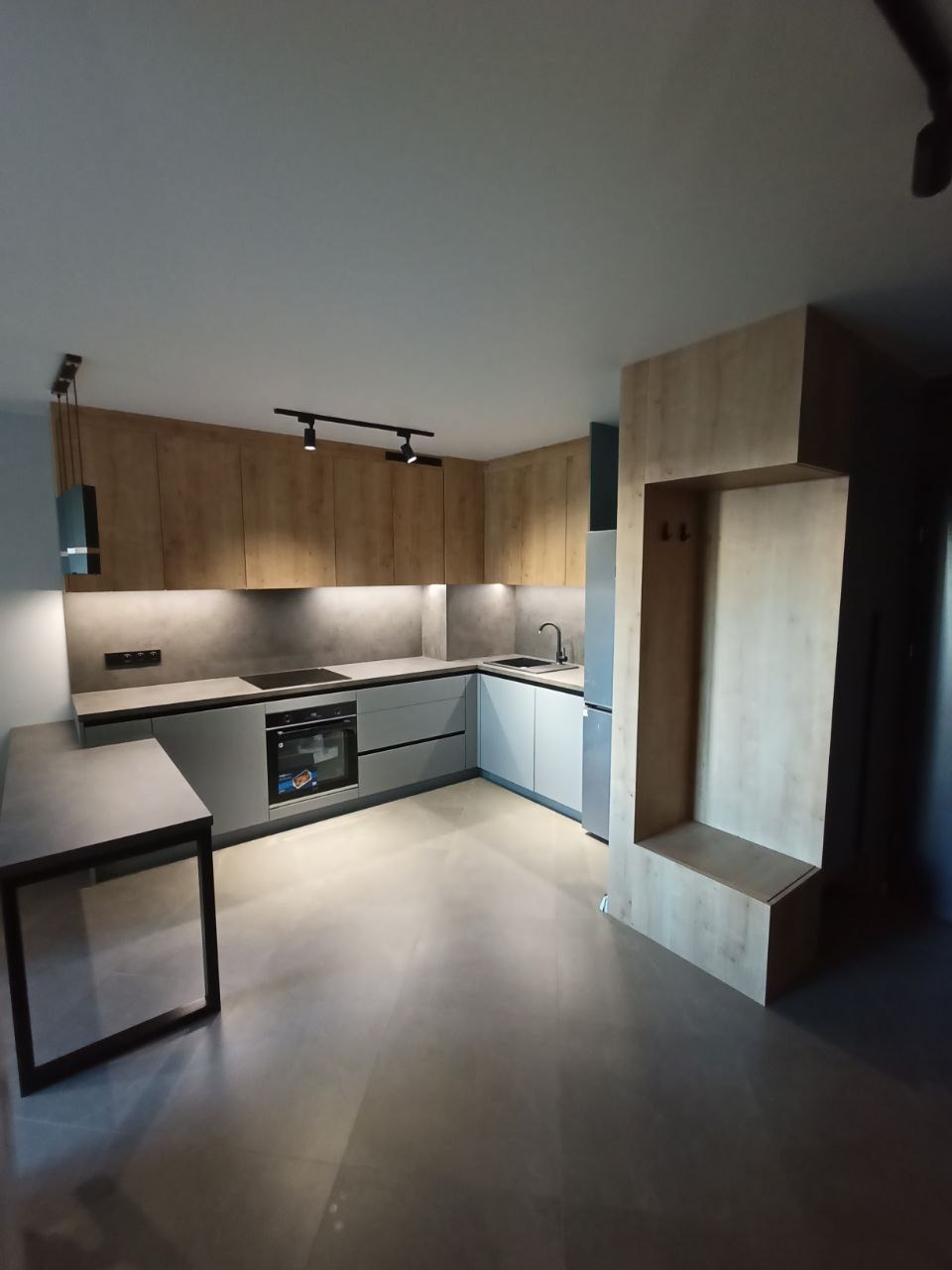 Nowoczesna kuchnia w kształcie litery U z drewnianymi szafkami górnymi i szarymi dolnymi, oświetleniem punktowym i stołem z metalową podstawą. Minimalistyczny design.