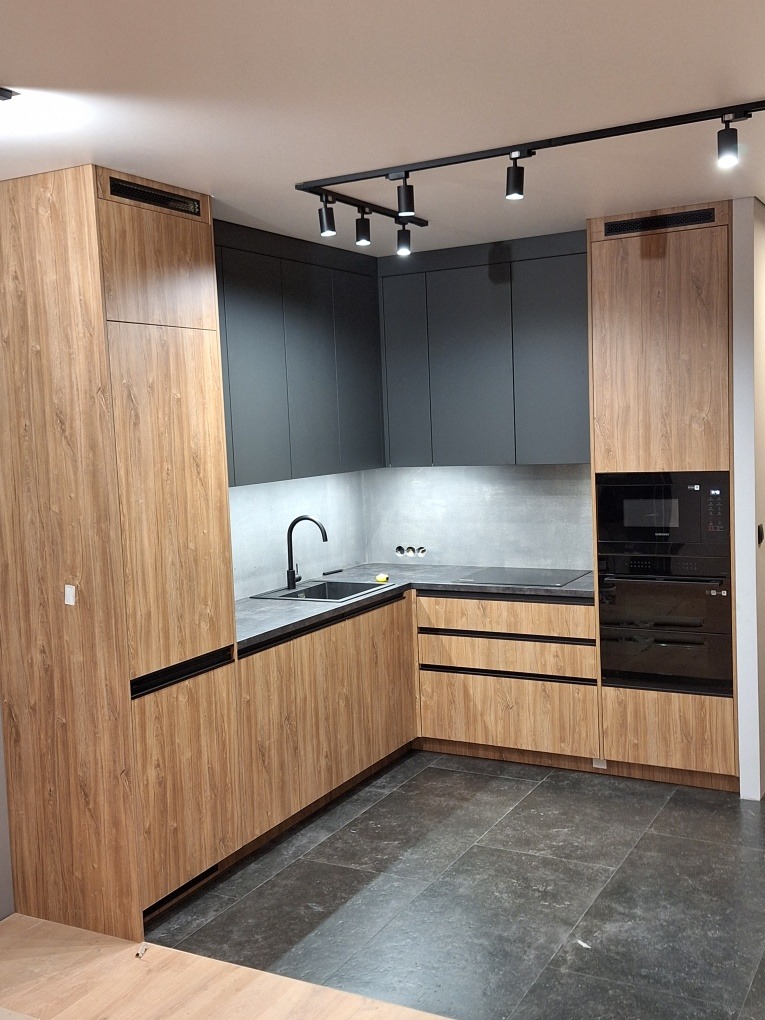 Nowoczesna kuchnia w odcieniach drewna i szarości, z czarnymi akcentami oświetlenia i sprzętów. Minimalistyczny design, widoczne detale wykończenia i oświetlenie punktowe.