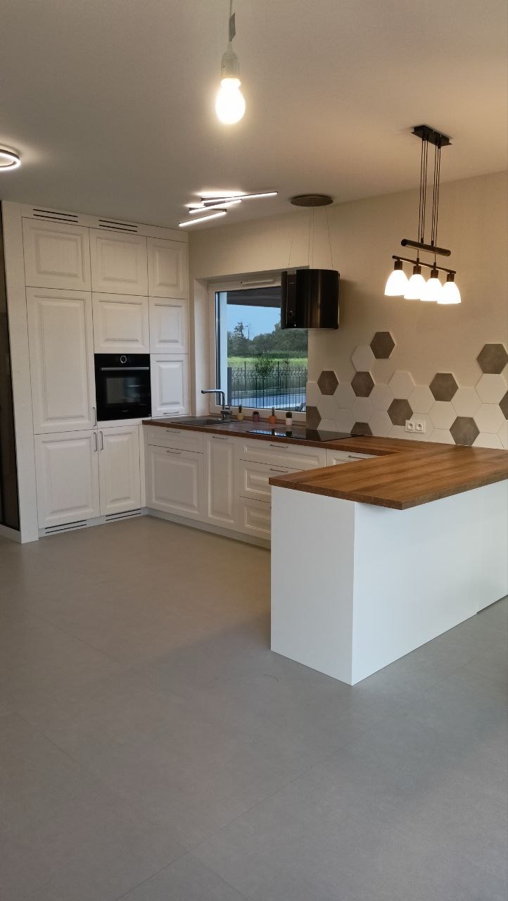 Jasna kuchnia z białymi szafkami, drewnianym blatem i heksagonalnymi płytkami na ścianie. Nowoczesne oświetlenie sufitowe. Widok na zewnątrz przez okno.