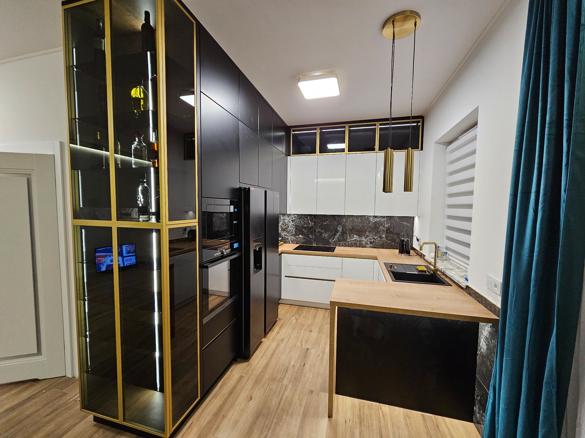 Nowoczesna kuchnia z czarnymi frontami i złotymi akcentami. Wysoka witryna z podświetleniem, drewniany blat i marmurowy backsplash. Minimalistyczne lampy wiszące.