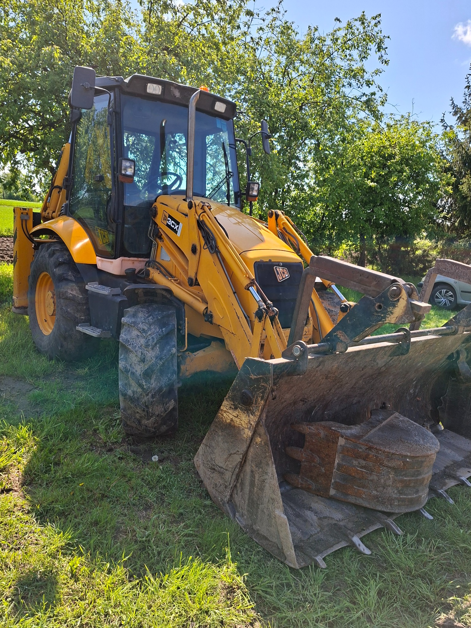 Żółta koparko-ładowarka JCB 3CX na trawiastym terenie, z widocznym łyżką i kabiną, w tle drzewa i fragment samochodu. Sprzęt gotowy do pracy.