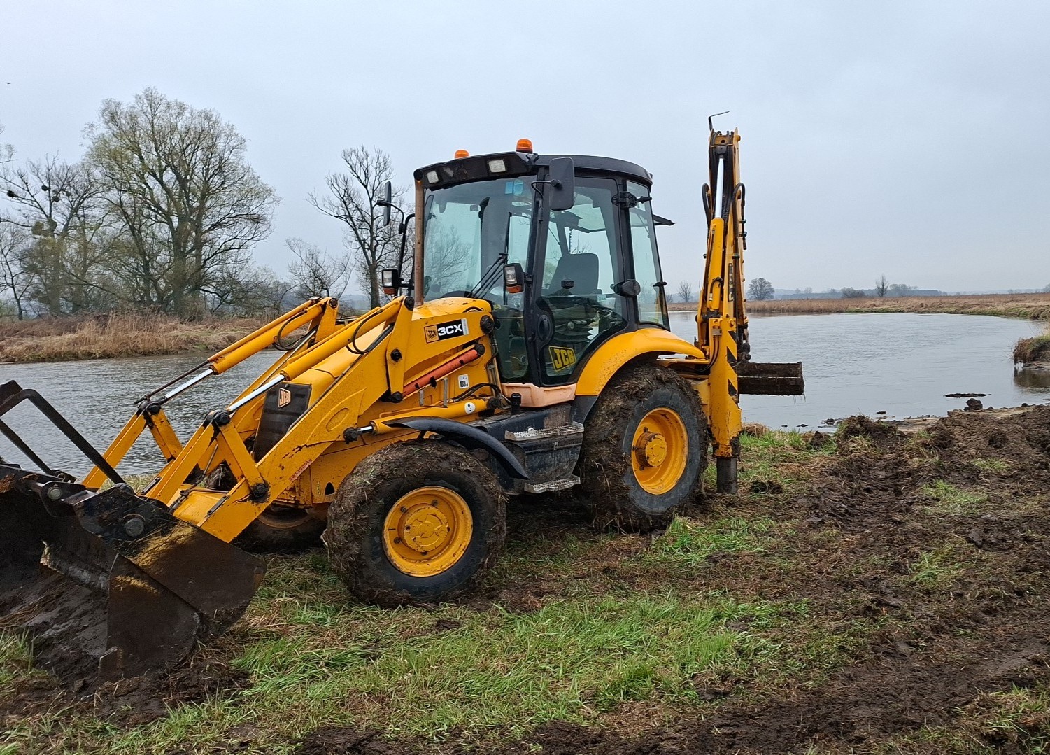 Żółta koparko-ładowarka JCB 3CX na błotnistym terenie nad brzegiem rzeki, z widocznym wykopem. Pochmurne niebo w tle. Maszyna gotowa do pracy ziemnych.