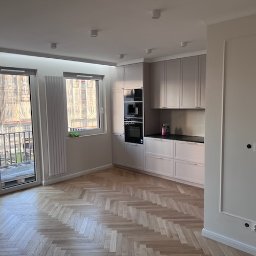 jednoosobowa działalność - Jasne wnętrze po remoncie: parkiet w jodełkę, zabudowa kuchenna w odcieniach beżu i szarości, duże okna z widokiem na budowę. Minimalistyczny styl, gładkie ściany.