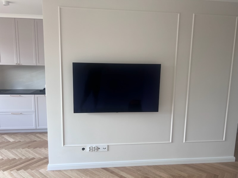 Telewizor na ścianie z dekoracyjnymi listwami w jasnym salonie z parkietem w jodełkę i widoczną kuchnią w tle. Nowoczesny design.