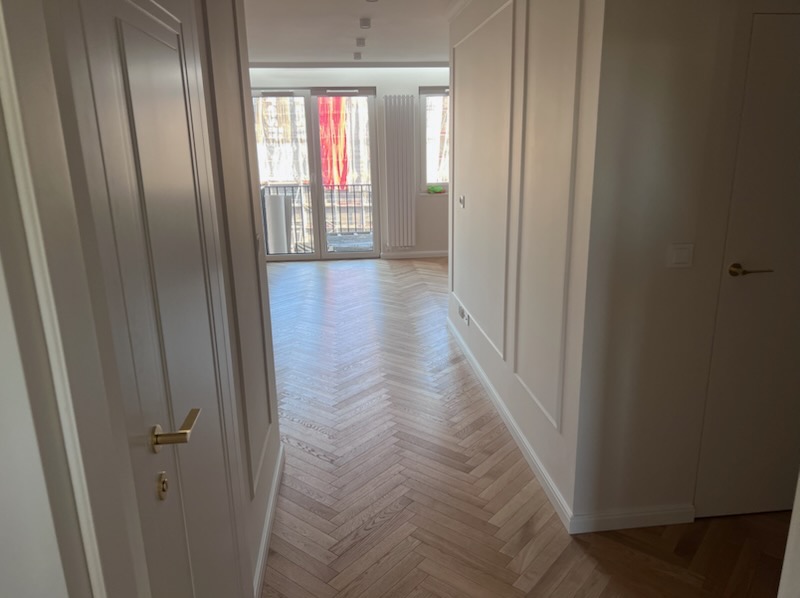 Jasne parkiety ułożone w jodełkę w odnowionym wnętrzu mieszkania, widok z korytarza przez otwarte drzwi na balkon. Ściany w kolorze złamanej bieli z dekoracyjnymi listwami.