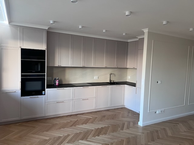 Jasna kuchnia z zabudową w odcieniach szarości i bieli, czarnym blatem i parkietem w jodełkę. Widoczne oświetlenie sufitowe i dekoracyjne listwy na ścianie.