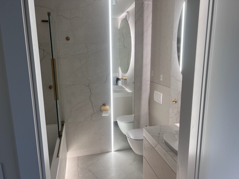 Elegancka łazienka z marmurowymi płytkami, oświetleniem LED i złotymi akcentami. Widok zza drzwi na wannę z parawanem i toaletę.