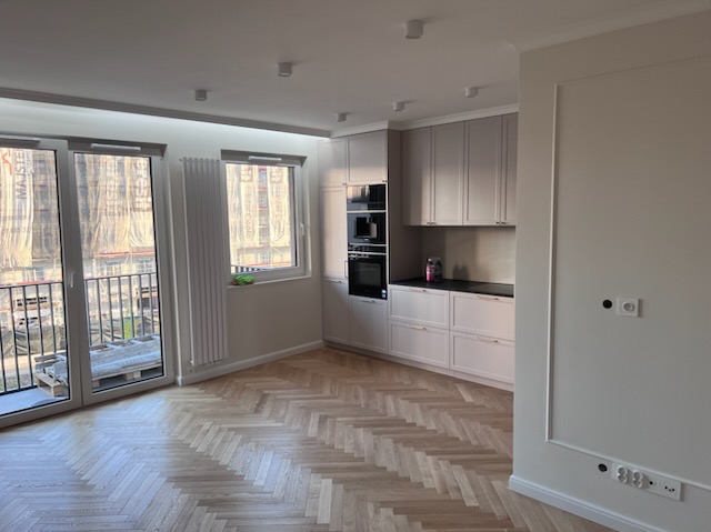 Jasne wnętrze po remoncie: parkiet w jodełkę, zabudowa kuchenna w odcieniach beżu i szarości, duże okna z widokiem na budowę. Minimalistyczny styl, gładkie ściany.