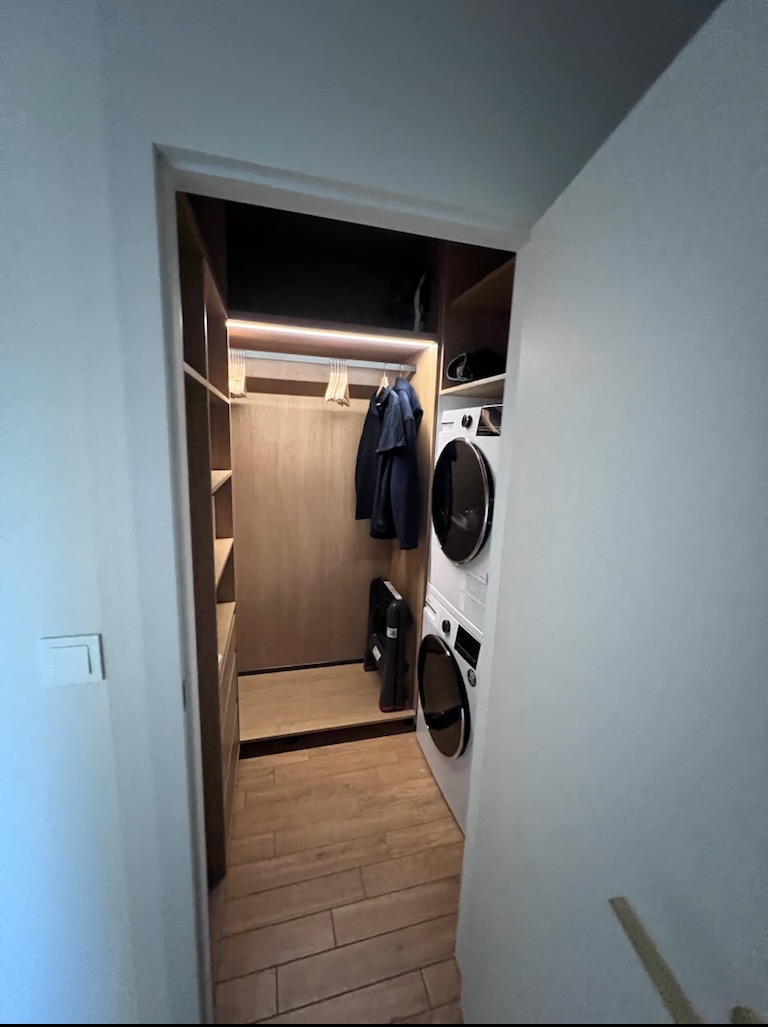 Funkcjonalna garderoba z pralką i suszarką w zabudowie, oświetlenie LED, drewniane półki i podłoga. Ubrania wiszą na wieszakach.