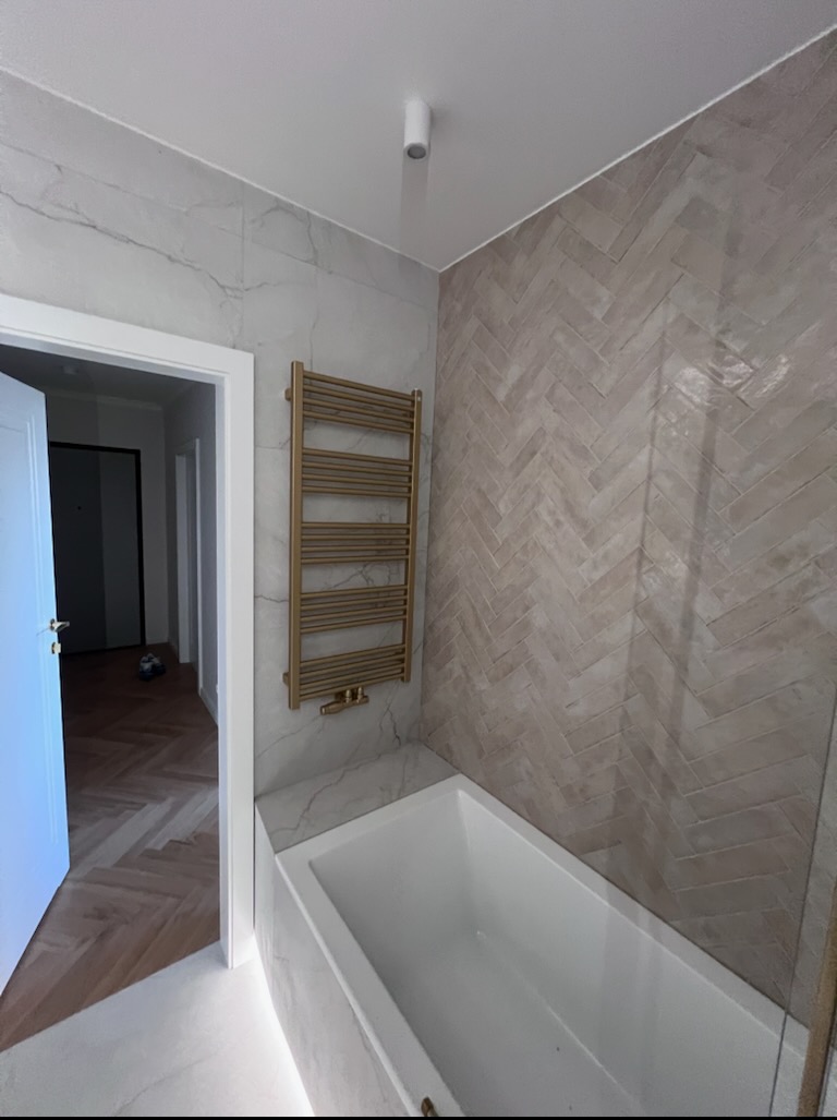 Wykończona łazienka z beżową mozaiką na ścianie ułożoną w jodełkę, białą wanną i złotym grzejnikiem drabinkowym. Widoczny fragment podłogi z parkietem w jodełkę.