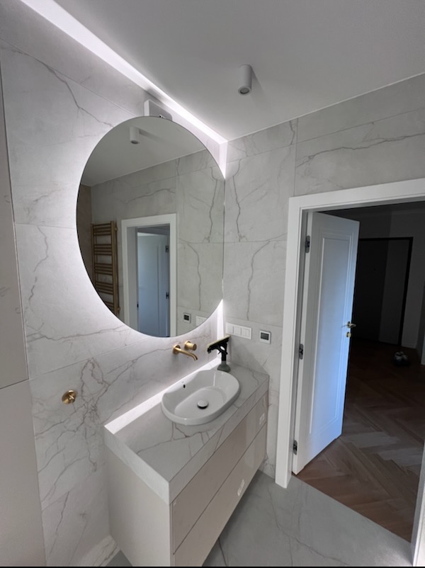 Nowoczesna łazienka z okrągłym lustrem z podświetleniem LED, marmurowymi płytkami i szafką pod umywalką w jasnych odcieniach. Widoczny fragment parkietu w sąsiednim pomieszczeniu.