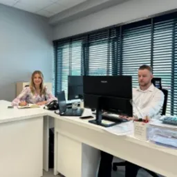 Biuro z jasnymi ścianami. Dwoje pracowników siedzi przy biurkach z monitorami. Na biurkach: laptopy, notatniki, długopisy. Żaluzje w oknach. Oświetlenie sufitowe.