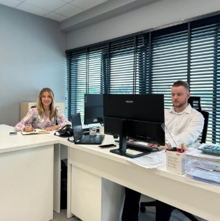 Biuro z jasnymi ścianami. Dwoje pracowników siedzi przy biurkach z monitorami. Na biurkach: laptopy, notatniki, długopisy. Żaluzje w oknach. Oświetlenie sufitowe.