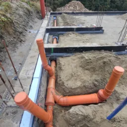 Pomarańczowe rury kanalizacyjne ułożone w wykopie, widoczne fragmenty fundamentów i izolacji, na tle ziemi i zieleni. Prace ziemne i instalacyjne w toku.