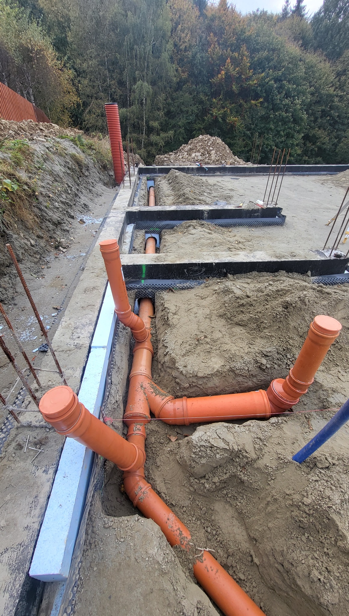 Pomarańczowe rury kanalizacyjne ułożone w wykopie, widoczne fragmenty fundamentów i izolacji, na tle ziemi i zieleni. Prace ziemne i instalacyjne w toku.