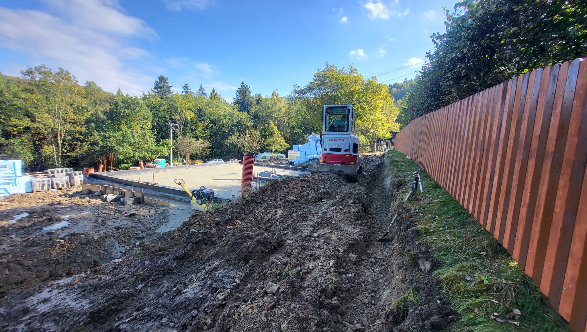 Fundament pod budowę domu jednorodzinnego z widocznym wykopem pod instalacje i mini koparką Takeuchi na tle zieleni i drewnianego ogrodzenia.