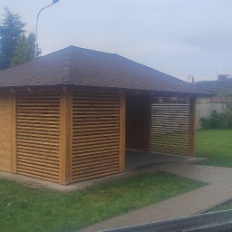 TMar-BUD - Drewniana altana z brązowym dachem na zielonym trawniku, z widocznymi deskami obok. Widok na szarą kostkę brukową i ogrodzenie w tle.