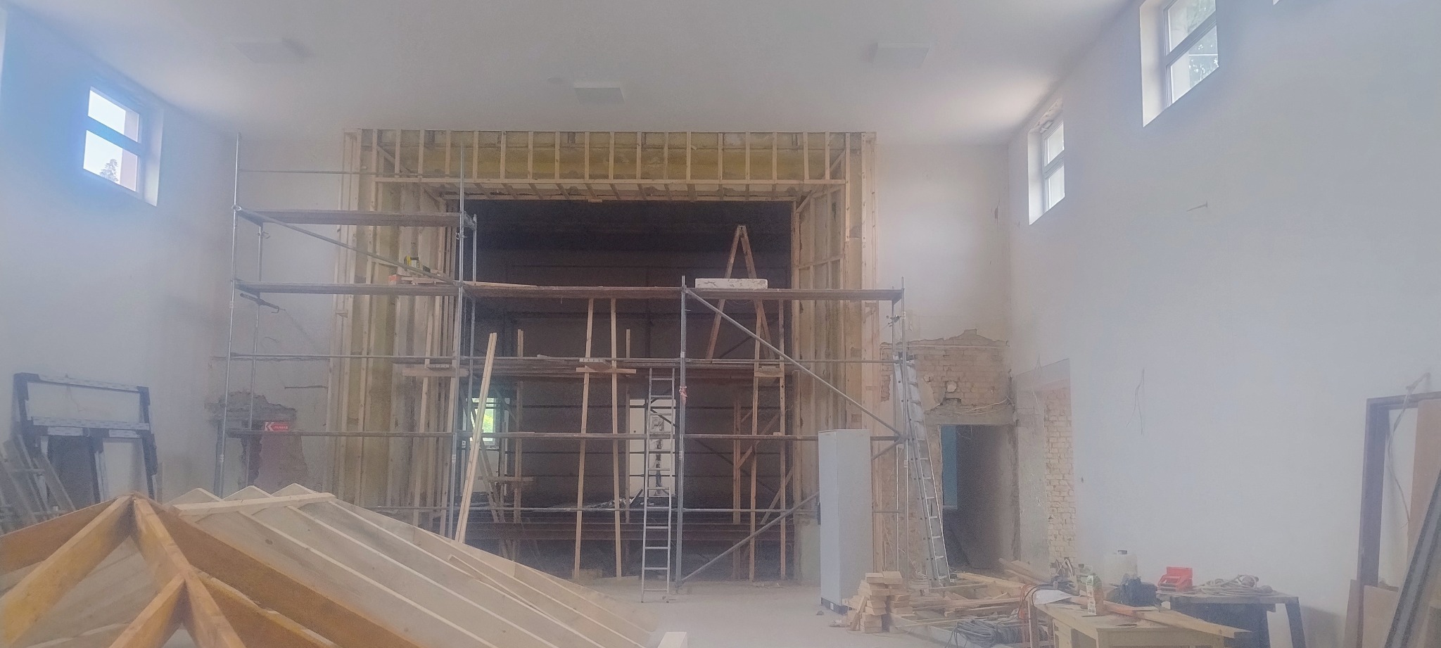 Wnętrze w trakcie remontu: drewniana konstrukcja ściany z izolacją, rusztowania i częściowo odsłonięta cegła. Widoczne okna w górnej części ściany.