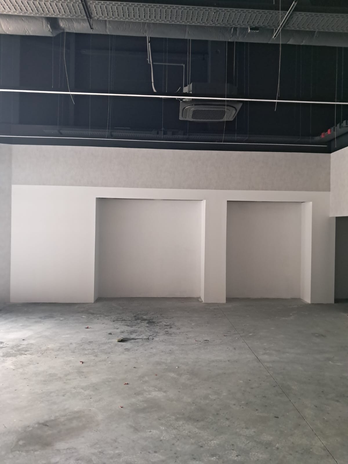 Surowe wnętrze z wnękami w ścianie, szara tapeta w górnej części, sufit z instalacjami. Podłoga betonowa, widoczne ślady po pracach budowlanych.