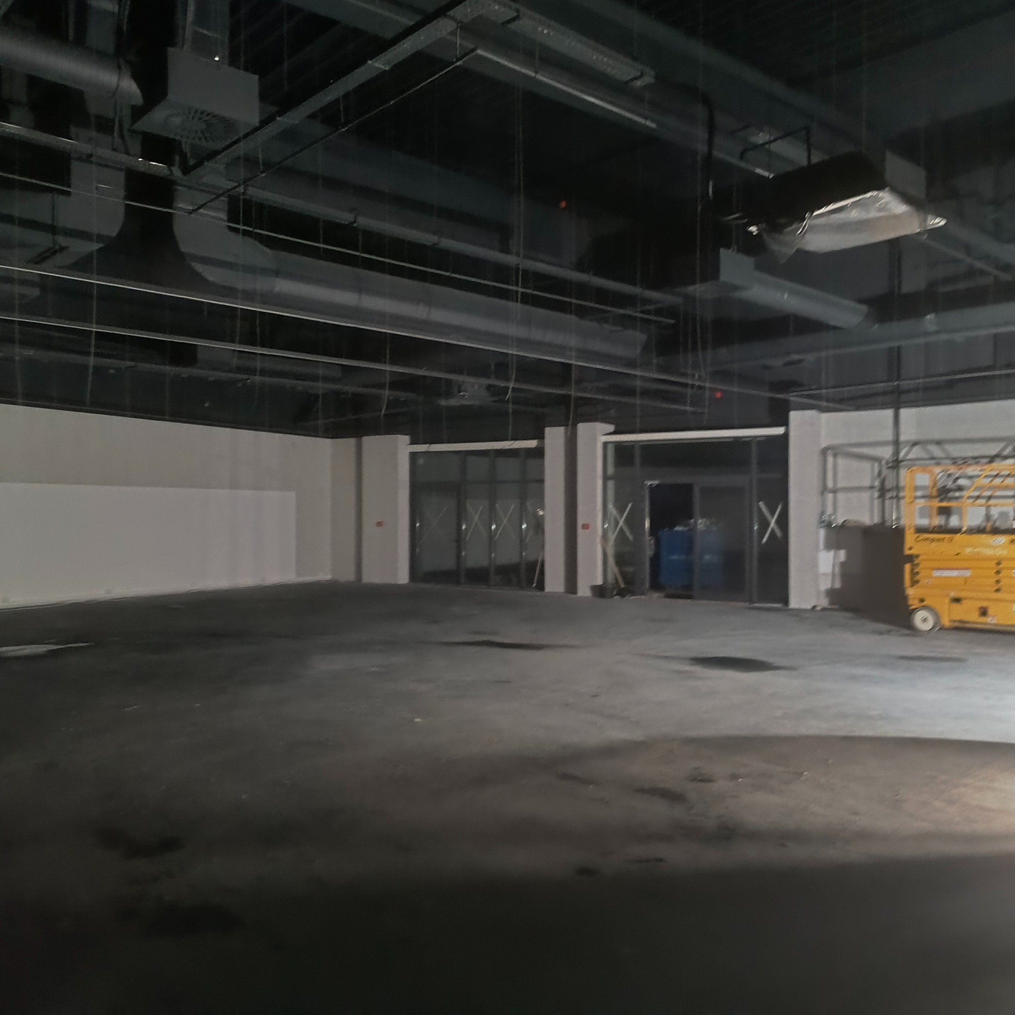 Wnętrze hali w trakcie prac wykończeniowych. Widoczne ściany, sufit z instalacjami, podłoga z plamami oraz podnośnik nożycowy. Częściowo zamontowane drzwi.