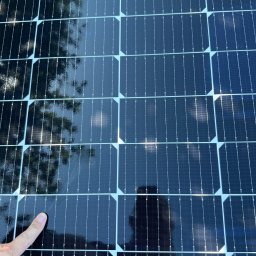 PPHU Eco&Clean Grzegorz Blat - Błyszczące panele fotowoltaiczne z odbiciem drzew i postaci, wskazane palcem. Układ paneli w regularnej siatce, widoczne detale konstrukcyjne.