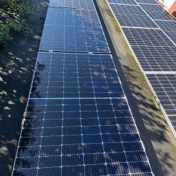 PPHU Eco&Clean Grzegorz Blat - Rząd paneli fotowoltaicznych na dachu, widoczne mocowania i cień drzewa. Błyszcząca powierzchnia paneli odbija niebo. Fragment dachu pokryty papą.