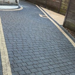 PPHU Eco&Clean Grzegorz Blat - Ułożona, grafitowa kostka brukowa z jasnymi pasami oddzielającymi, prowadząca do drewnianej bramy i domu z zieloną elewacją. Widok z góry, słoneczny dzień.