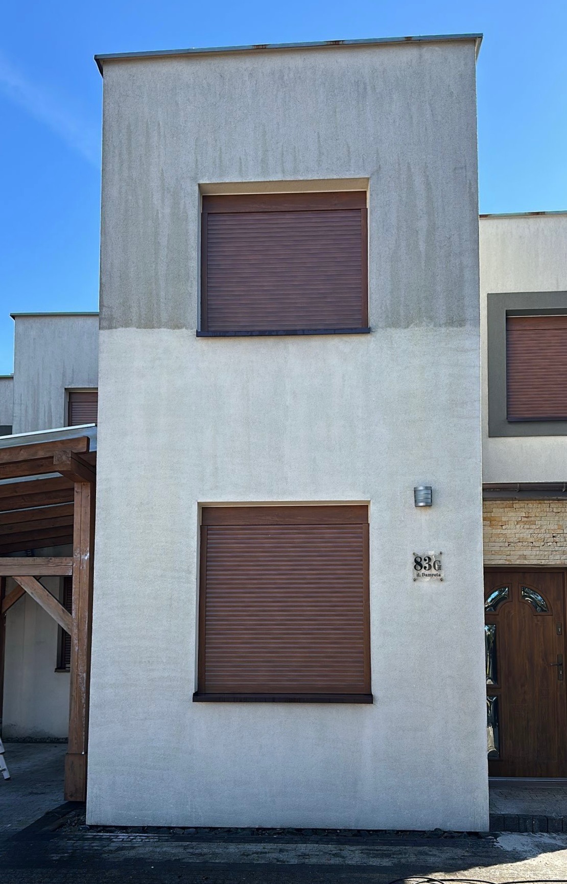 Dwukondygnacyjny budynek z szarą elewacją, brązowymi roletami okiennymi i numerem domu 83G. Górna część elewacji jest wyraźnie brudniejsza od dolnej.