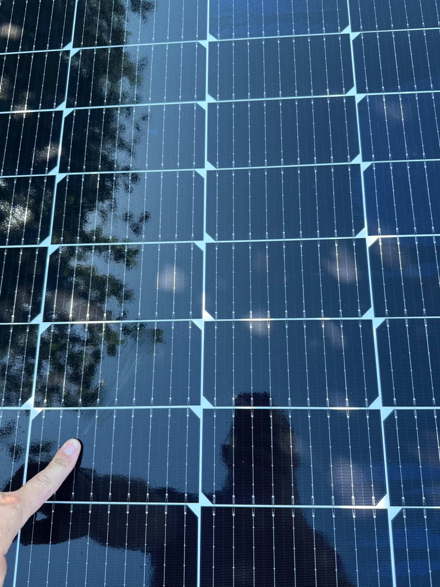 Błyszczące panele fotowoltaiczne z odbiciem drzew i postaci, wskazane palcem. Układ paneli w regularnej siatce, widoczne detale konstrukcyjne.