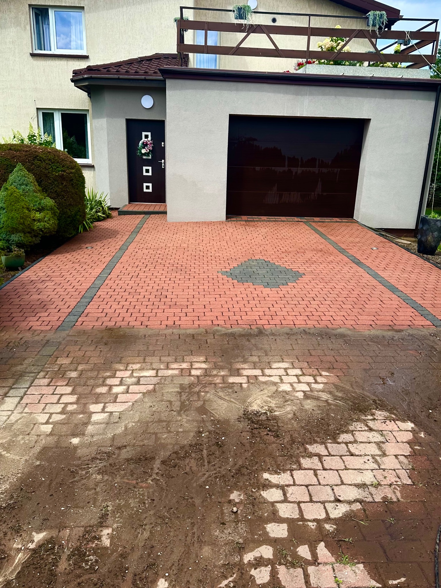 Częściowo umyta kostka brukowa na podjeździe przed domem jednorodzinnym. Widoczna różnica w czystości kostki, od brudnej po czystą, z wzorem z szarej kostki.