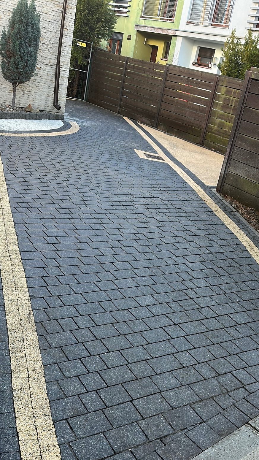 Ułożona, grafitowa kostka brukowa z jasnymi pasami oddzielającymi, prowadząca do drewnianej bramy i domu z zieloną elewacją. Widok z góry, słoneczny dzień.