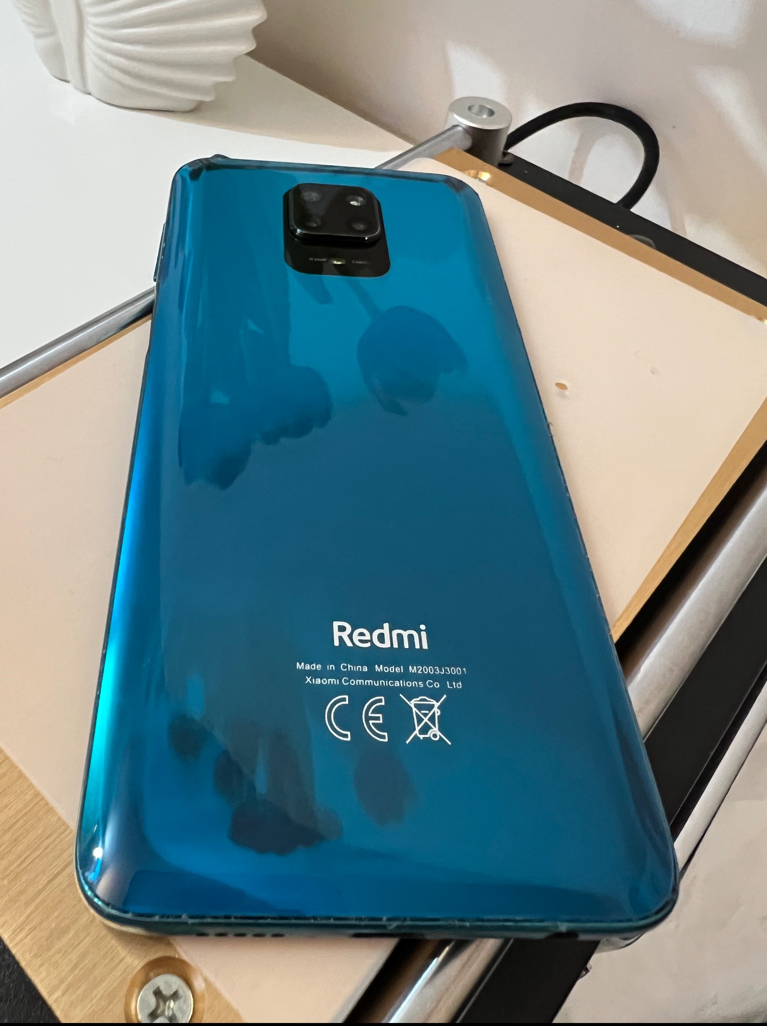 Błyszczący, turkusowy smartfon Redmi z widocznym aparatem i oznaczeniami producenta leży na jasnym blacie. Widoczne odbicia światła na obudowie.