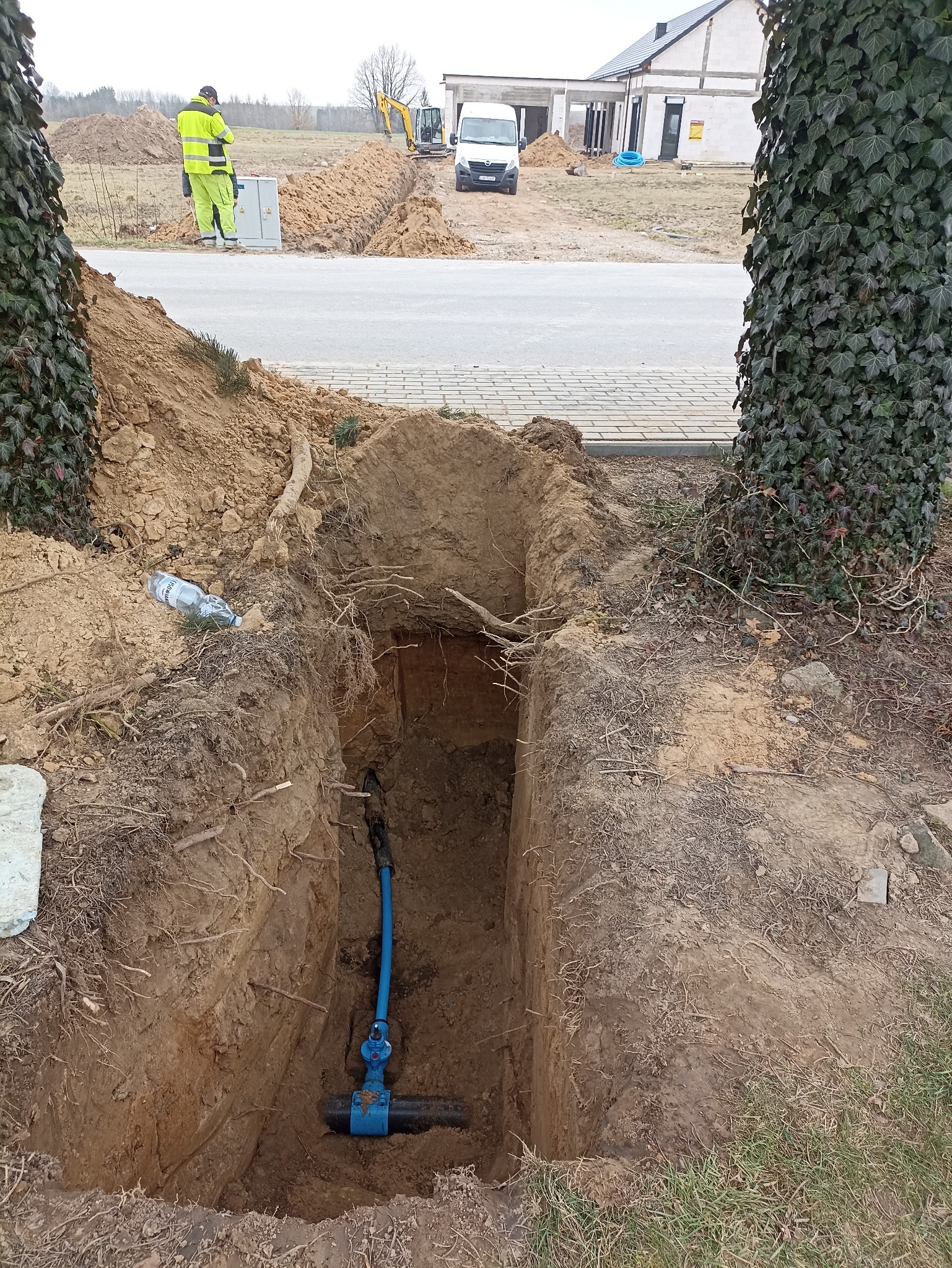 Głęboki wykop z niebieską rurą i zaworem na dnie, widoczne korzenie. W tle robotnicy i wykopany rów w ziemi. Prace ziemne przy budowie domu.