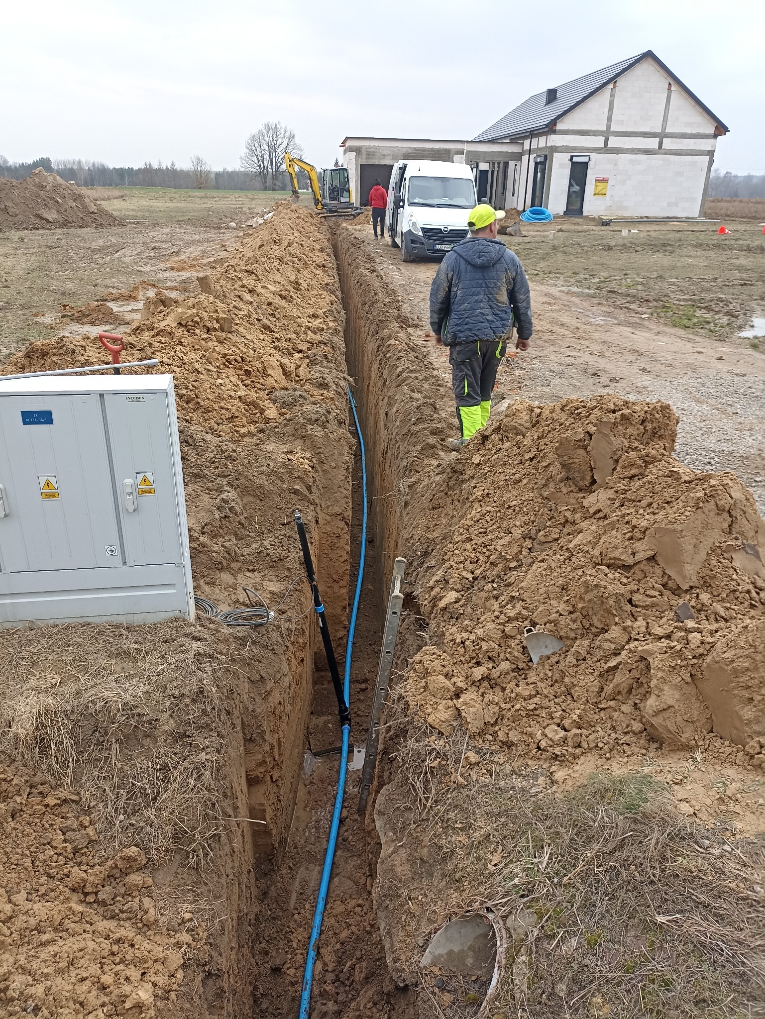 Wykop z ułożoną niebieską rurą, widoczna skrzynka elektryczna, koparka i pracownicy w tle. Prace ziemne na placu budowy domu jednorodzinnego.