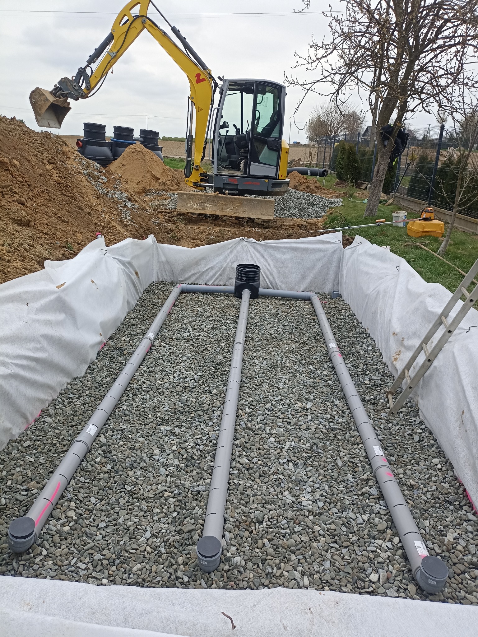 Instalacja drenażowa w wykopie wyłożonym geowłókniną, z rurami drenarskimi i warstwą żwiru. W tle koparka i zbiorniki.