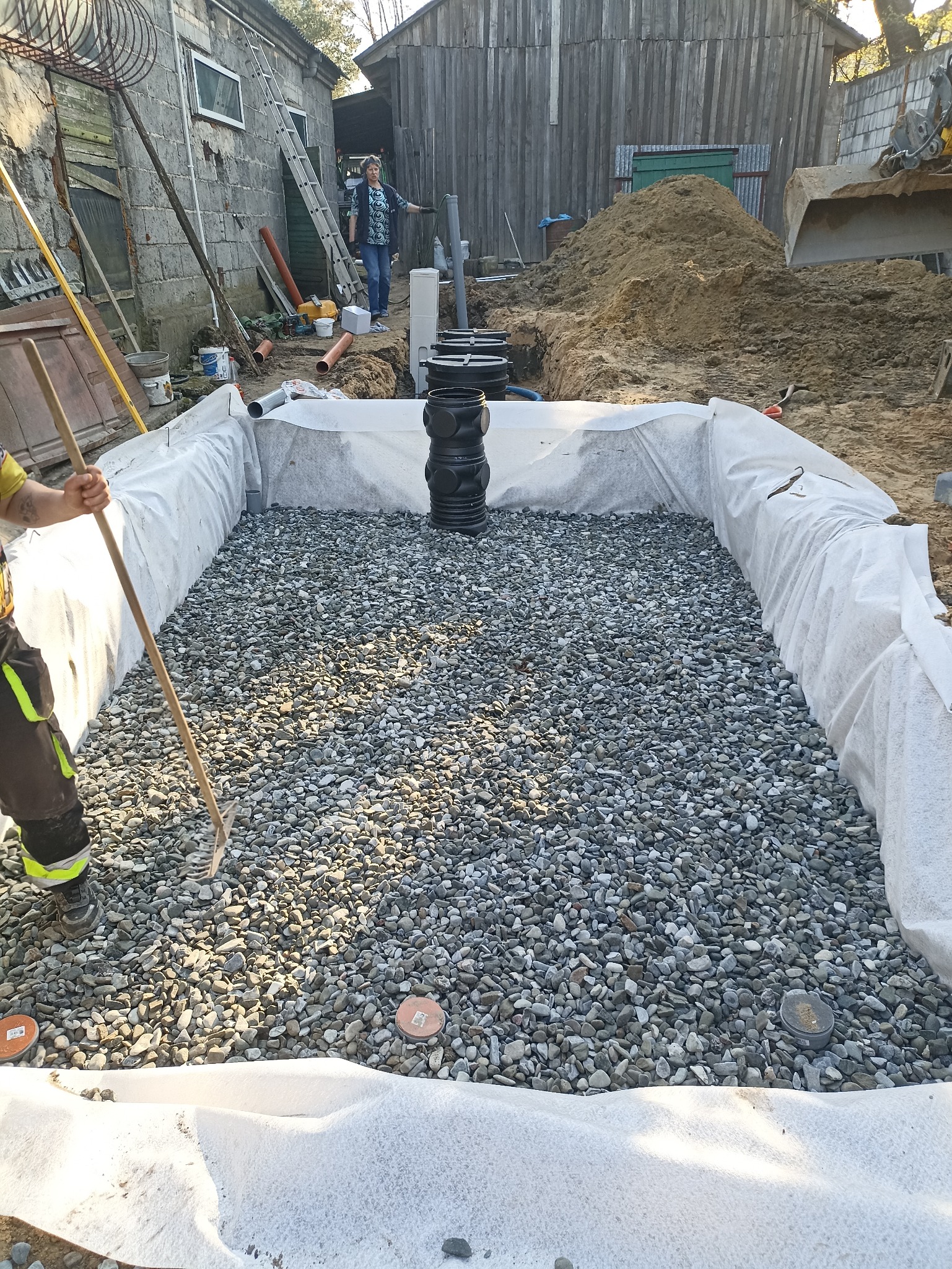 Instalacja drenażowa w wykopie wyłożonym geowłókniną, wypełnionym żwirem. W tle prace ziemne i budynki gospodarcze, osoba nadzorująca.