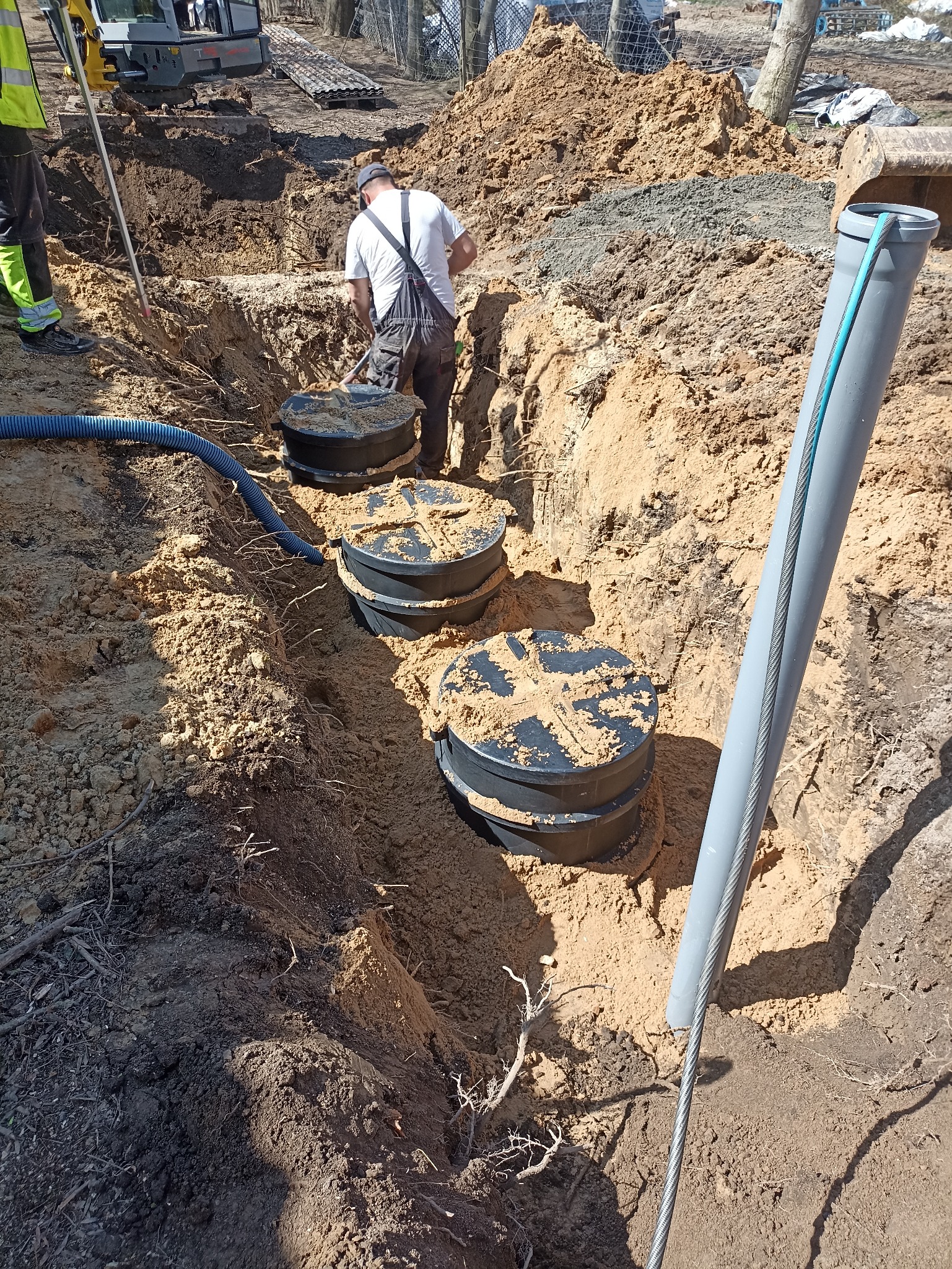 Montaż czarnych zbiorników w wykopie, rury i pracownik w tle. Widoczne elementy instalacji i ziemia. Prace ziemne w toku, dzień słoneczny.