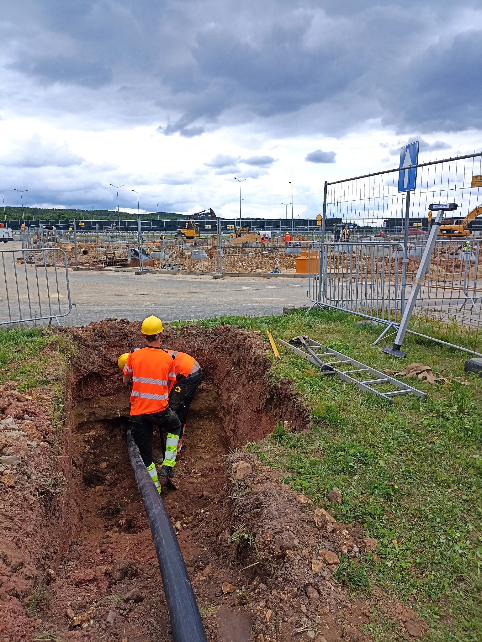 Dwóch robotników w wykopie układa czarną rurę na tle placu budowy z maszynami i pochmurnego nieba. Widoczne barierki i drabina.