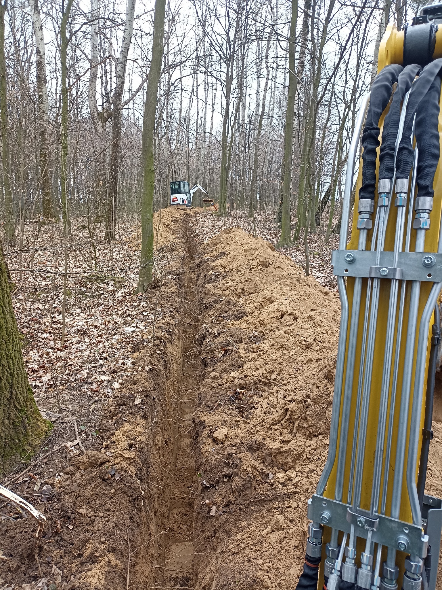 Wąski wykop w lesie, wykonany przez białą mini koparkę widoczną w tle. Na pierwszym planie fragment ramienia żółtej koparki.