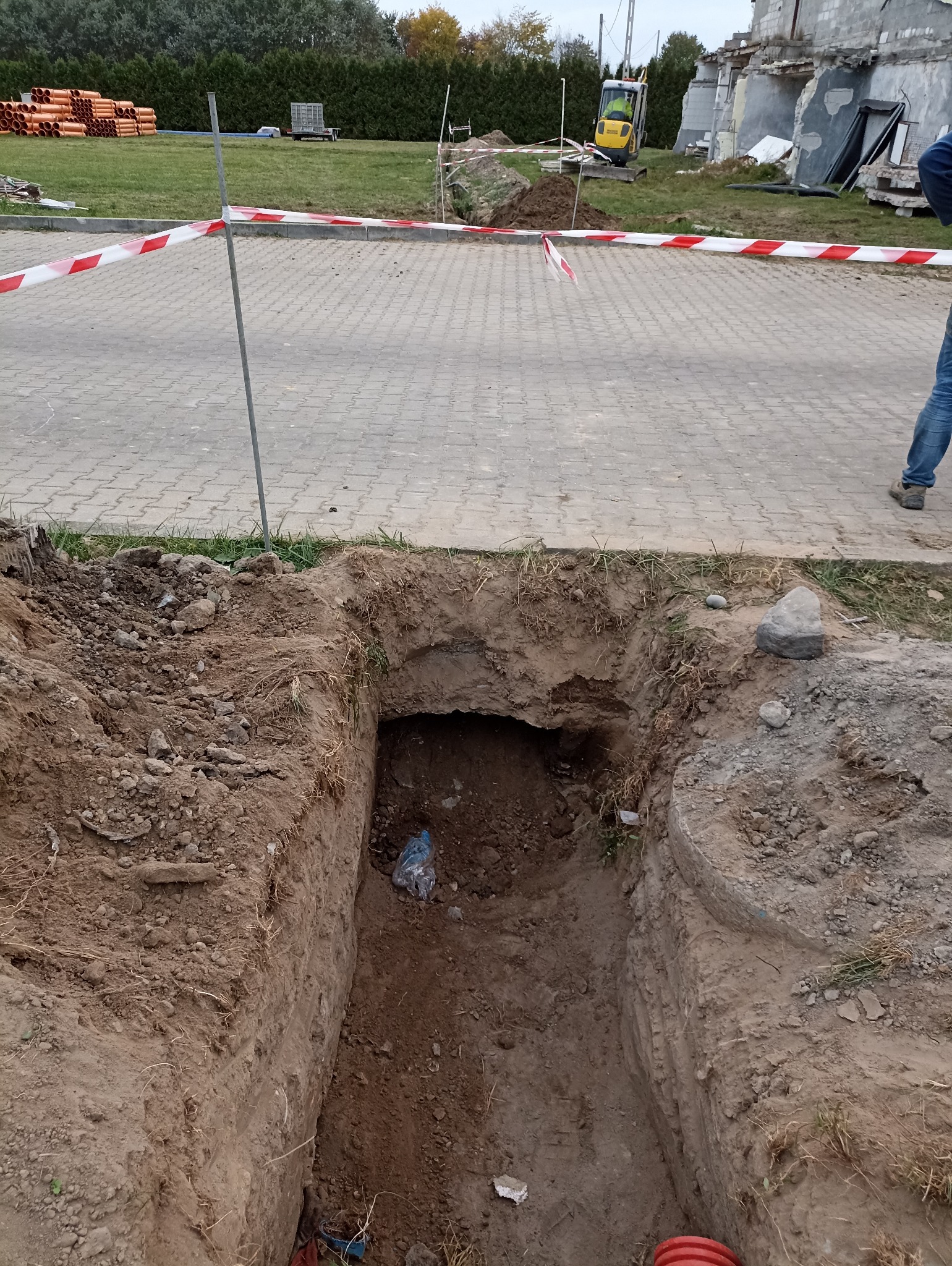 Głęboki wykop w ziemi, zabezpieczony taśmą ostrzegawczą. W tle widoczny minikoparka i sterta rur. Wykop pod instalacje.