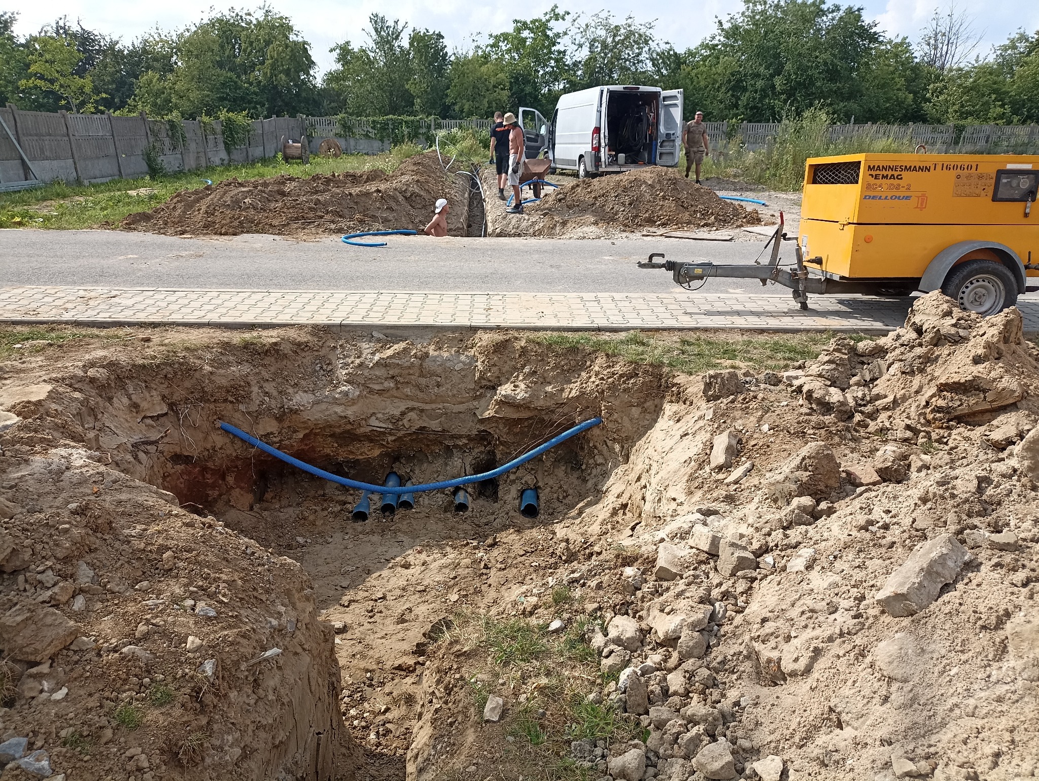 Głęboki wykop w ziemi z widocznymi niebieskimi rurami, w tle ekipa robotników przy białym samochodzie dostawczym i żółty agregat prądotwórczy na przyczepie.