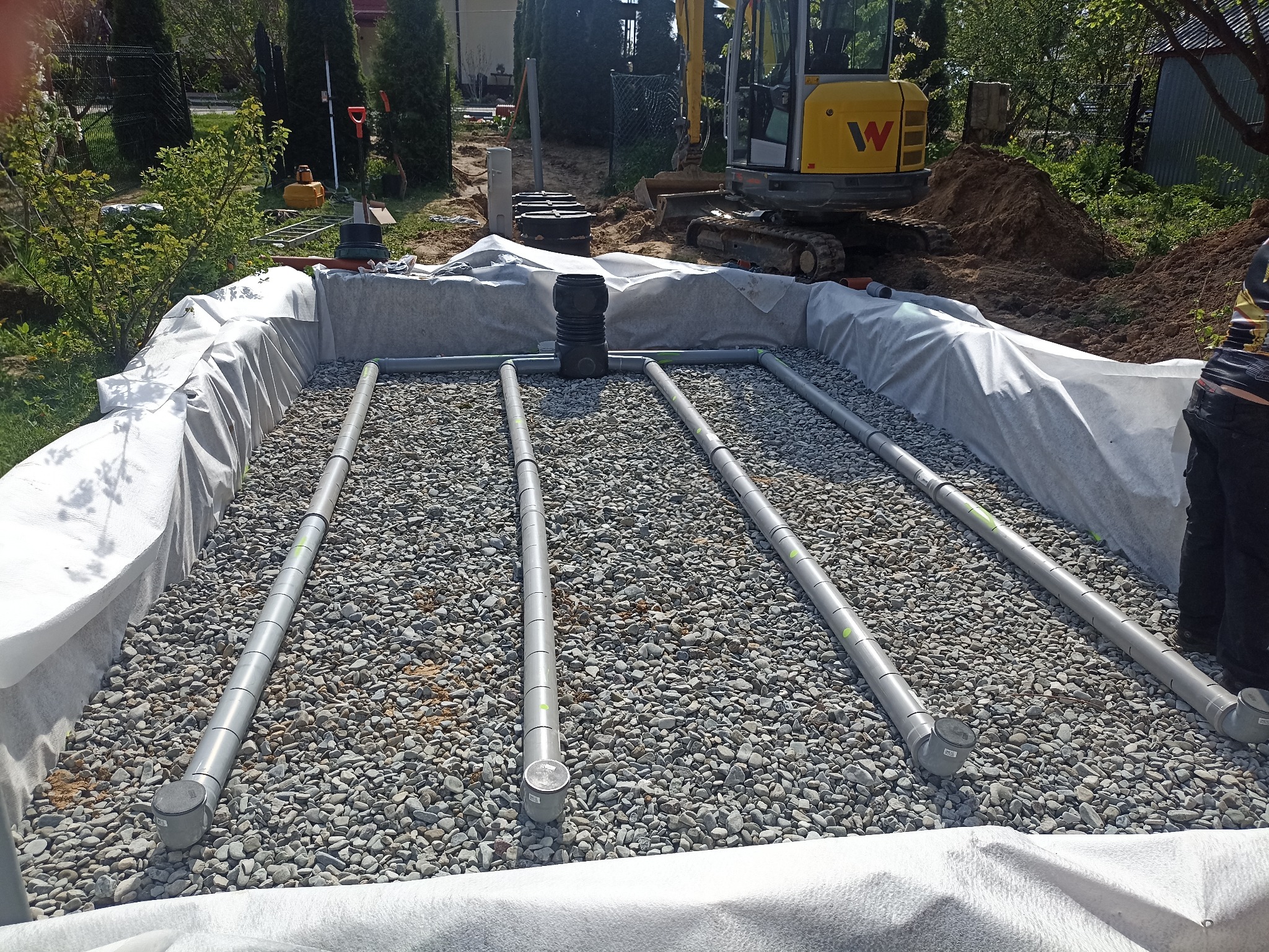 Drenaż rozsączający na żwirze, zabezpieczony białą geowłókniną w wykopie. W tle minikoparka i teren budowy.