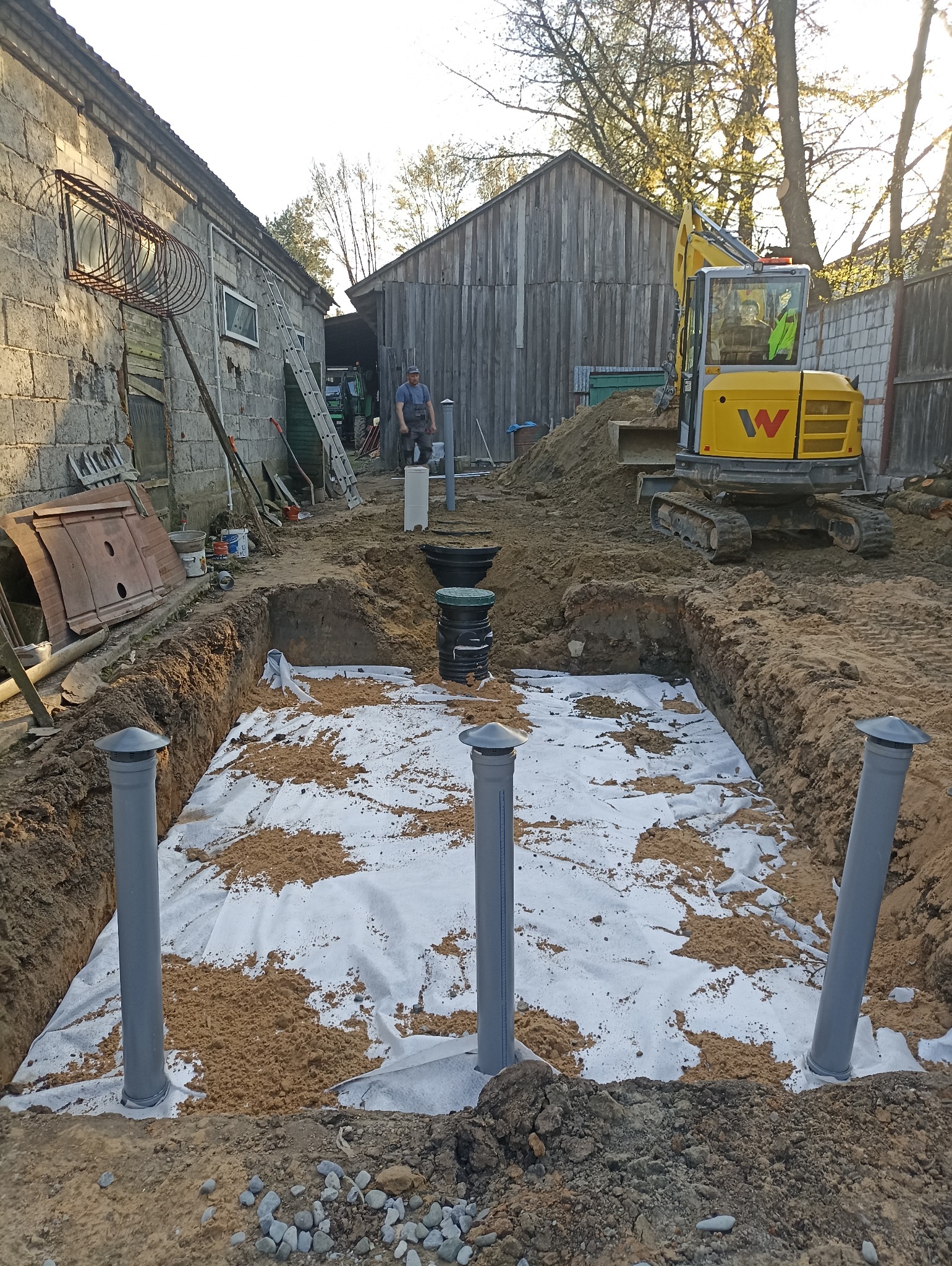 Wykop z geowłókniną i pionowymi rurami wentylacyjnymi, prawdopodobnie pod oczyszczalnię ścieków. W tle budynki gospodarcze i koparka.